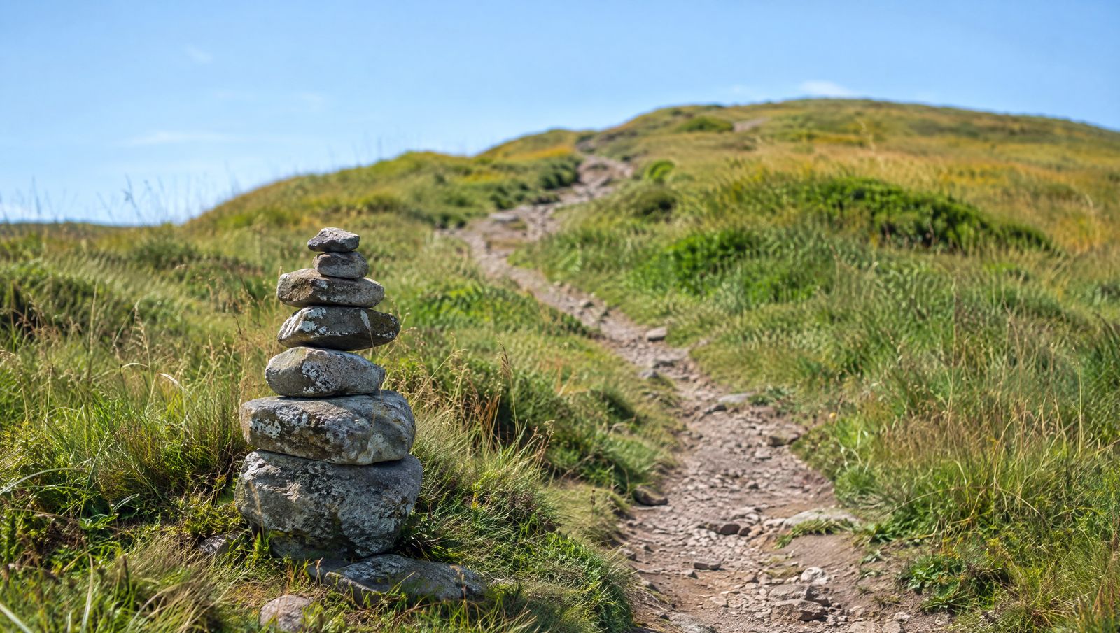 A small stone cairn beside a dirt trail on a green hillside with the path continuing uphill