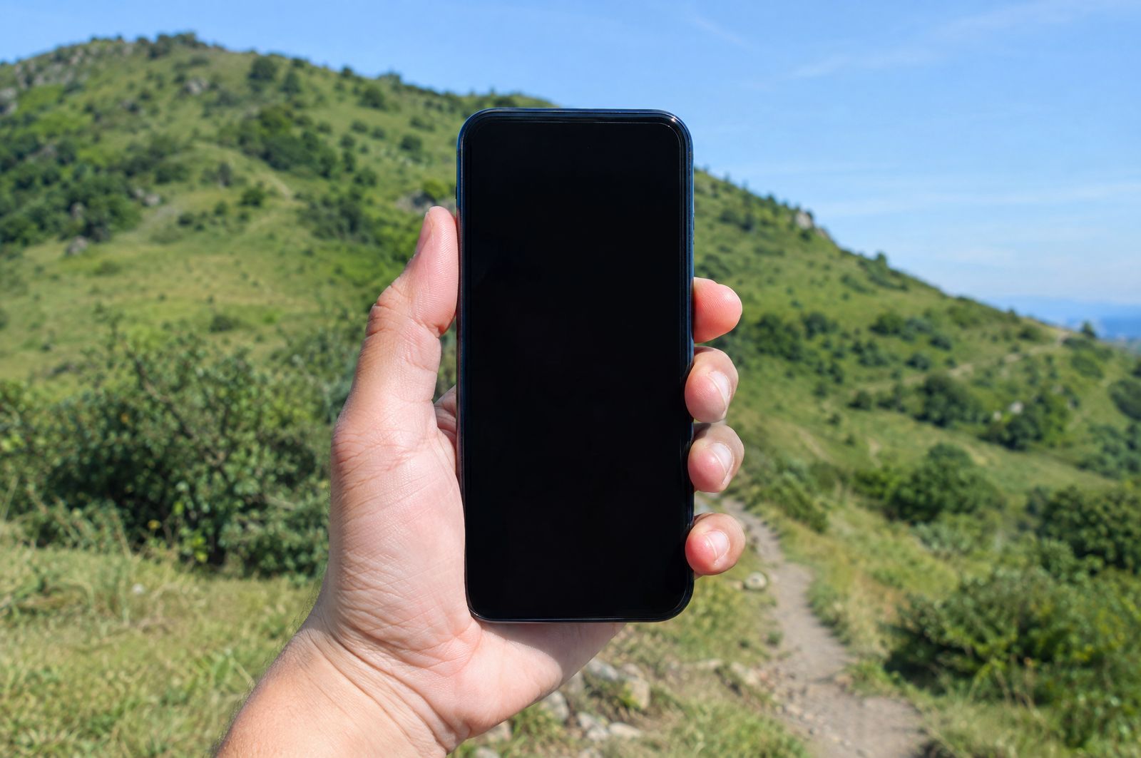 A hand holding a smartphone with a dark blank screen in front of a green hillside trail