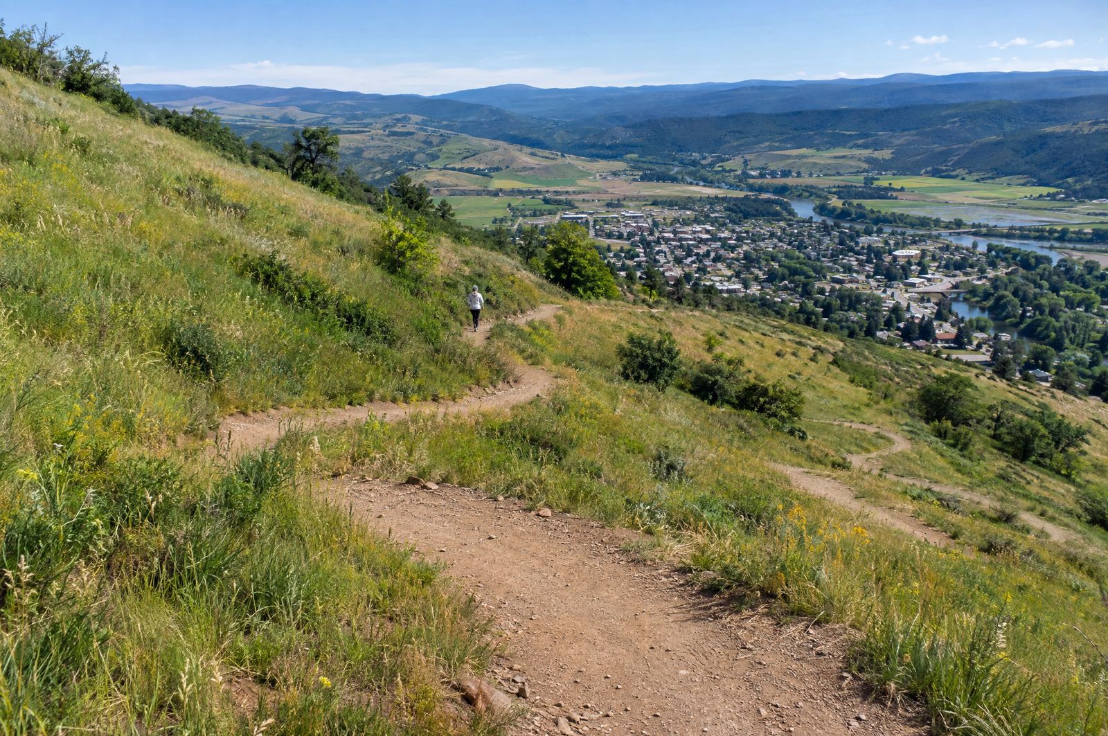 A dirt switchback trail zigzagging up a grassy hillside, one small distant walker seen from behind, a town in the valley below