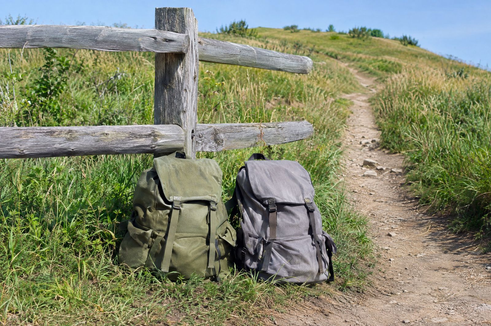 Two canvas daypacks leaning on a weathered wooden fence at a grassy trailhead, a dirt path rising behind